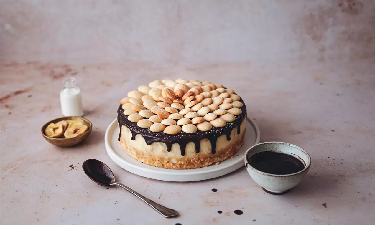 Kulatý cheesecake na bílé míse, zdobený vrstvou čokoládové polevy s kapkami po okrajích, na povrchu rovnoměrně rozložené banánové sušenky