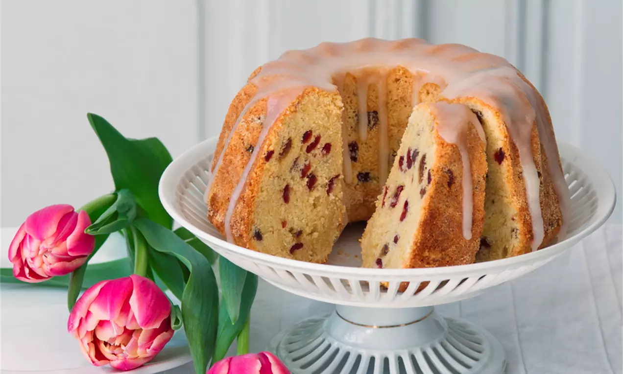 Mandlová bábovka s brusinkami světlé zlatavé barvy s lesklou citronovou polevou na bílé stojánku, vedle růžové tulipány na lněném ubruse