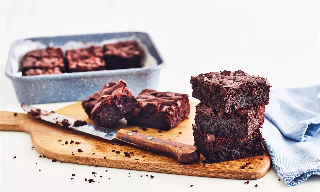 Čtvercové brownies s tmavou, bohatou texturou na dřevěném prkénku s nožem, vedle modré utěrky a zapékací mísy na bílém pozadí