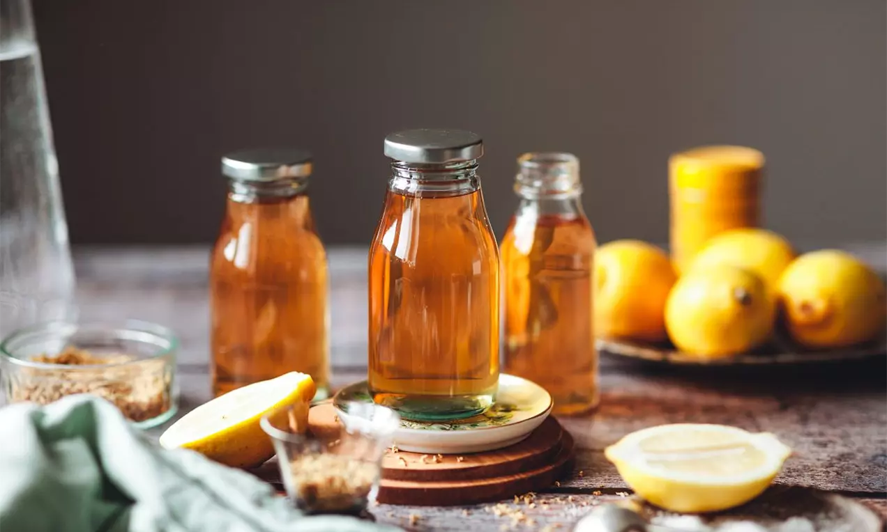 Bezový sirup v průhledných lahvích s kovovými víčky, jantarové barvy, na dřevěném stole vedle plátků citronu, celých citronů a skleničky cukru