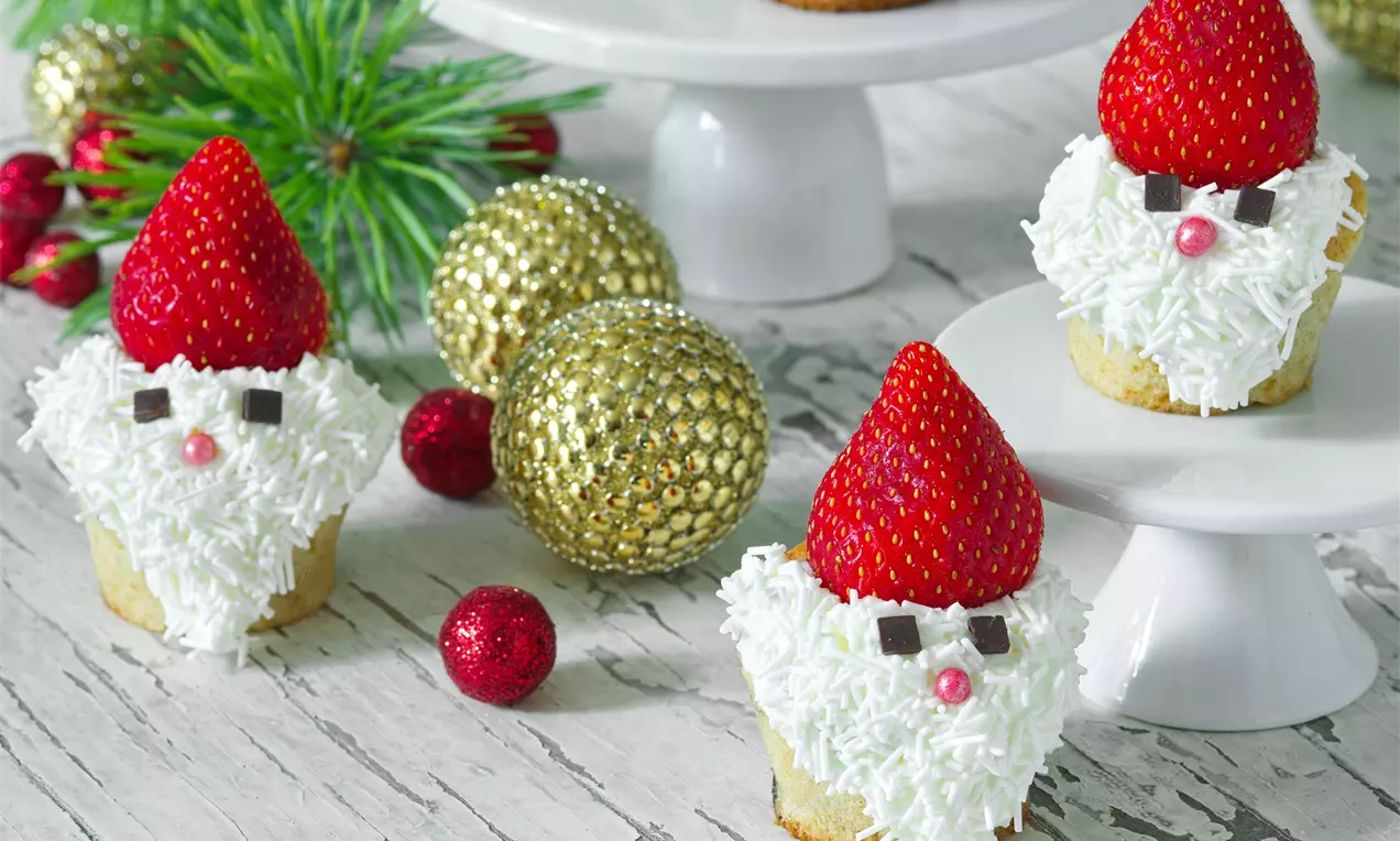 Muffiny se světlým korpusem zdobené bílým krémem a kokosem, červenými jahodami jako čepicemi, čokoládou a perličkami na dřevěném stole