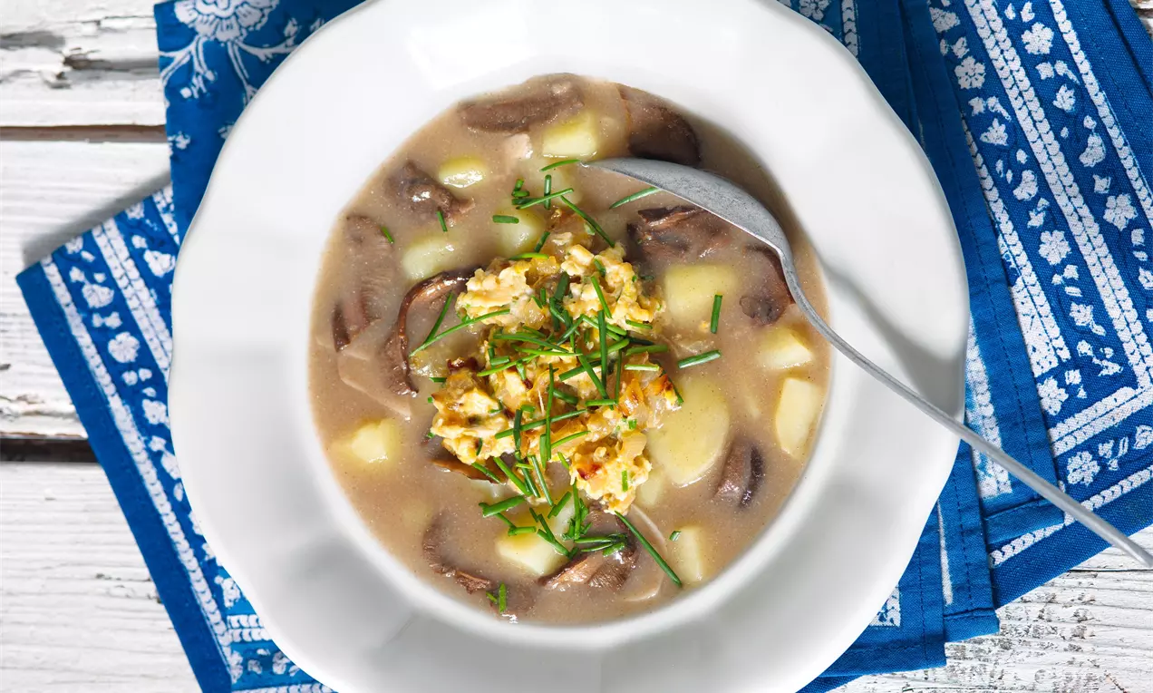Světle hnědá polévka s kousky hub, brambor a kopcem smažených vajec, posypaná nasekanou pažitkou na bílém talíři na modrém ubrousku