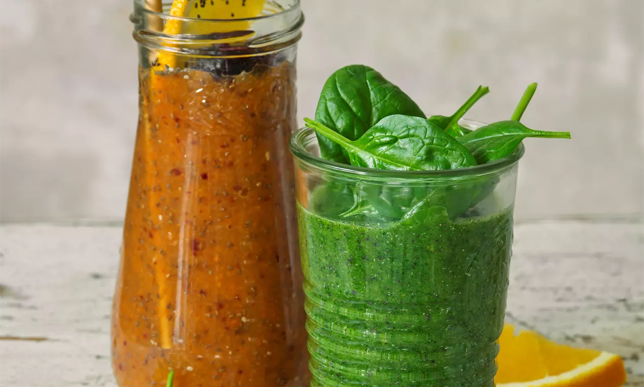 Hustý zelený smoothie ve sklenici zdobené čerstvými listy špenátu vedle oranžového smoothie v lahvi se slupkou pomeranče na dřevěném stole