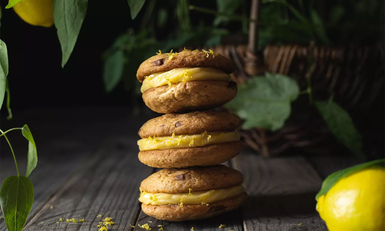 Světlé citronové cookies ve tvaru koleček spojené vanilkovým krémem, navrstvené do komínku na dřevěném stoje vedle citronů a lístků
