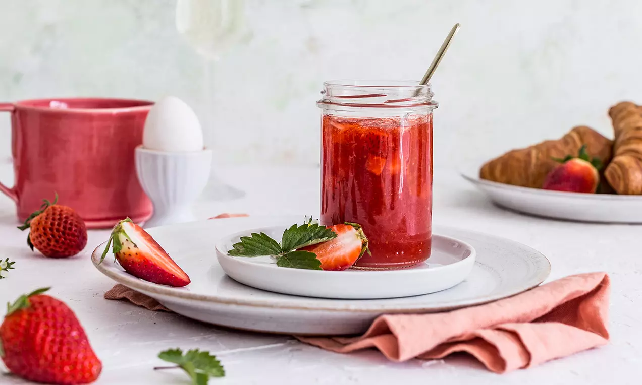Sklenice se sytě červeným jahodovým džemem s proseccem, usazená na bílém talíři, obklopená čerstvými jahodami, růžovou látkou a pečivem