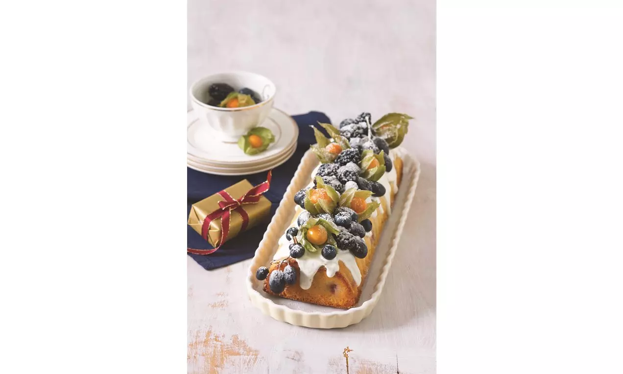 Obdélníkový hřbet zdobený bílou jogurtovou polevou, čerstvým hroznovým vínem, borůvkami, physalis a posypaný moučkovým cukrem na světlém podkladu
