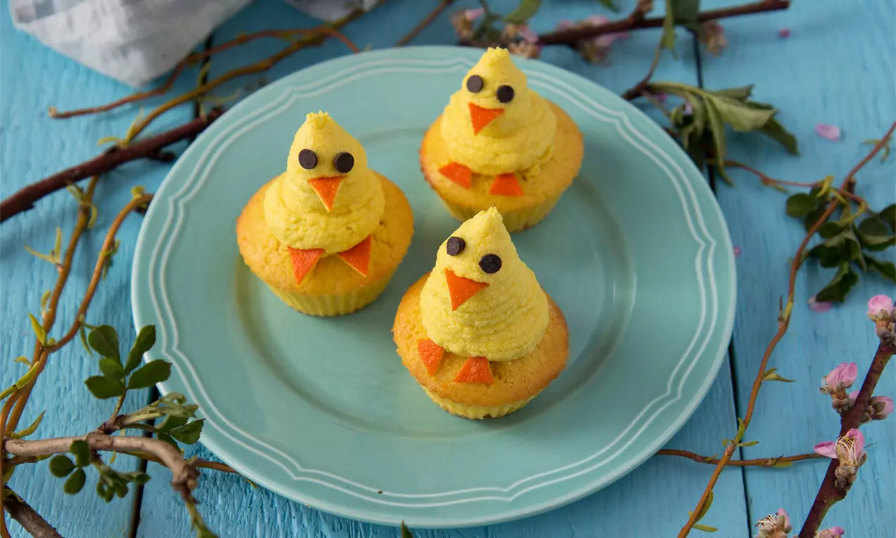 Velikonoční kuřátka z jemného mangového krému na žlutých cupcakes s čokoládovými očky, zdobená oranžovými zobáčky na světle modrém talíři