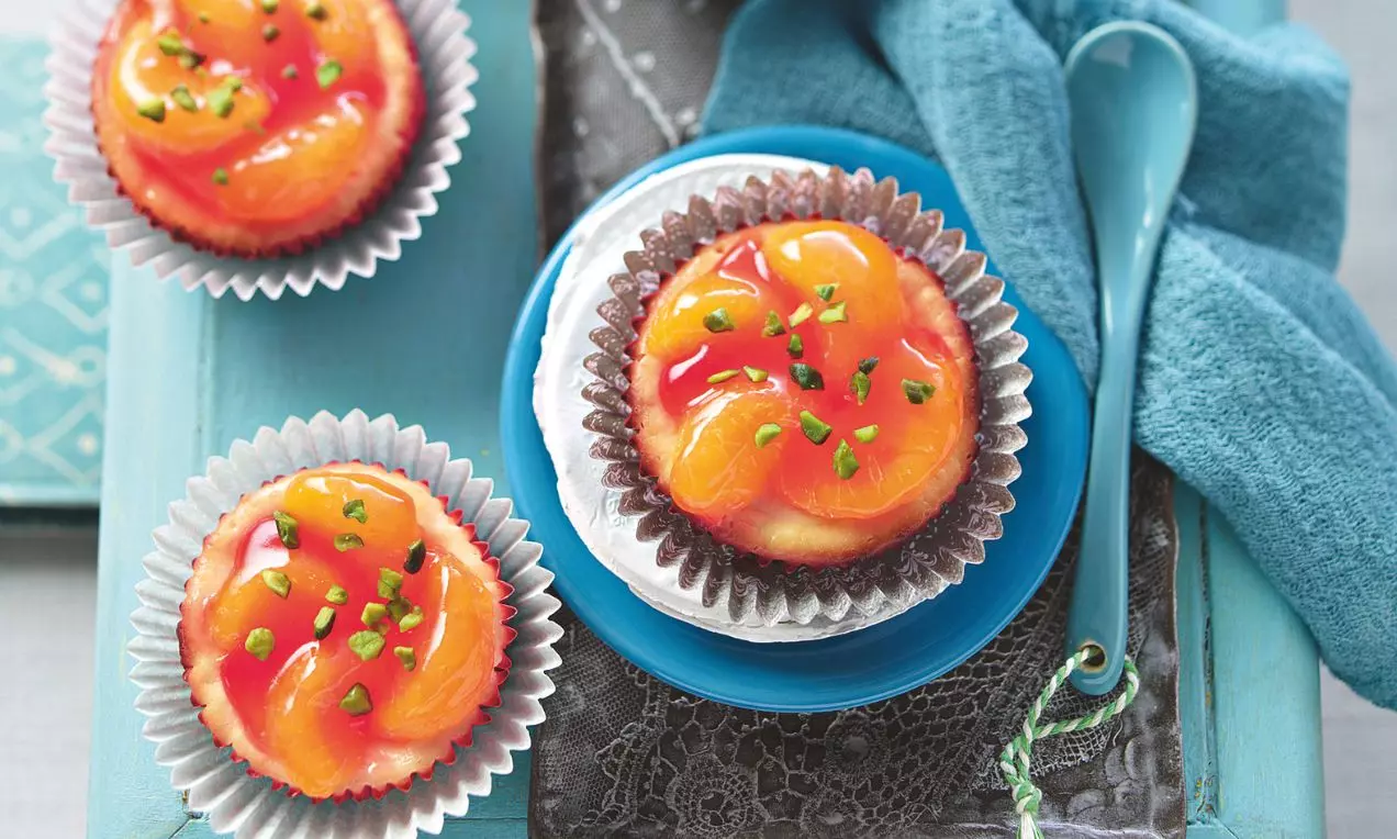 Mandarinkovo-tvarohové muffiny s lesklou oranžovou vrstvou mandarinek, jemným krémovým povrchem a drobnými zelenými ozdobami na modrém podkladu