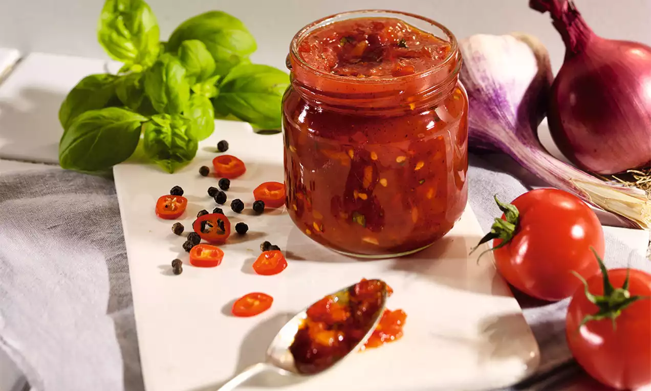 Sklenice s hustým červeným čatní plným drobných kousků rajčat a koření na světlém prkénku, obklopená čerstvými rajčaty, chilli a bazalkou