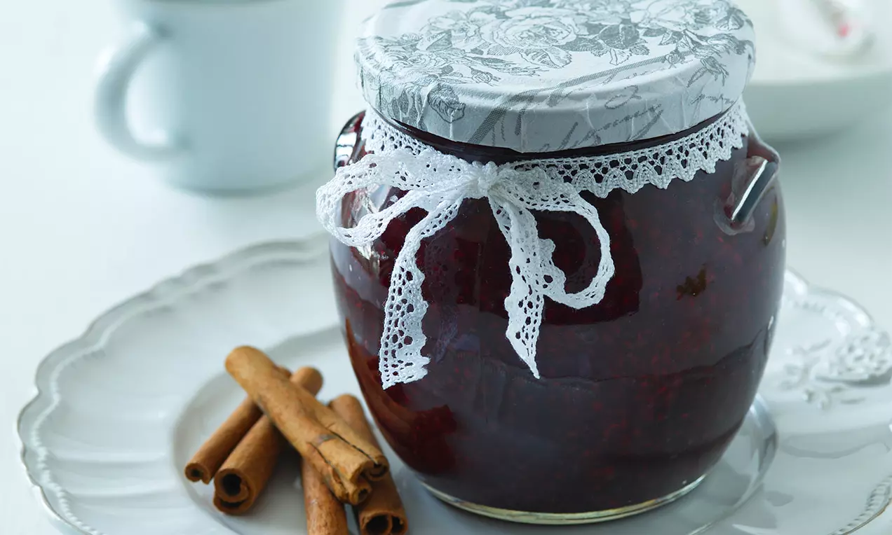 Sklenice s višňovou marmeládou tmavě červené barvy s krajkovým uzávěrem na bílém talíři vedle voňavých hnědých svitků skořice