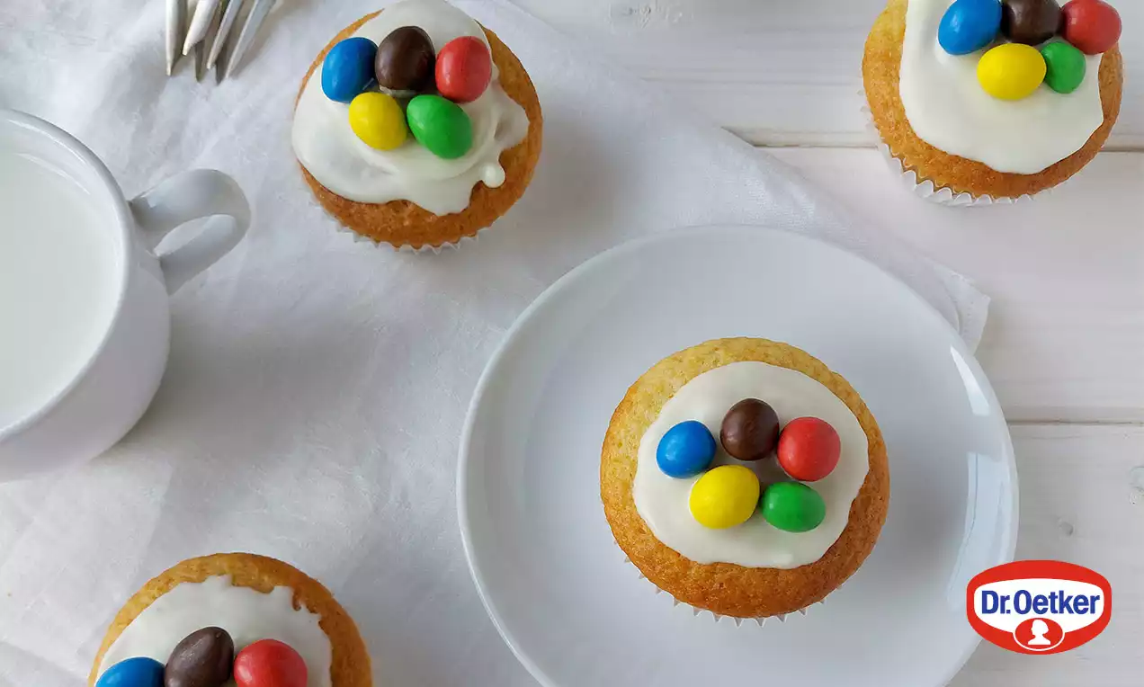 Zlatavé muffiny s bílou polevou, zdobené barevnými kulatými bonbony, na bílém talíři a látkovém ubrousku vedle šálku mléka a vidliček