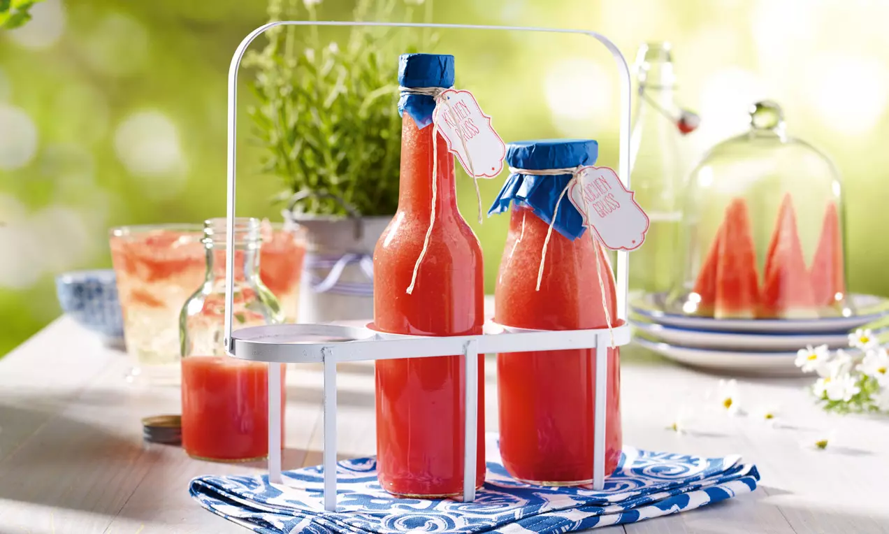 Červený melounový sirup ve štíhlých lahvích s modrými víčky a cedulkami, usazený v bílém stojanu na modrém látkovém ubrousku