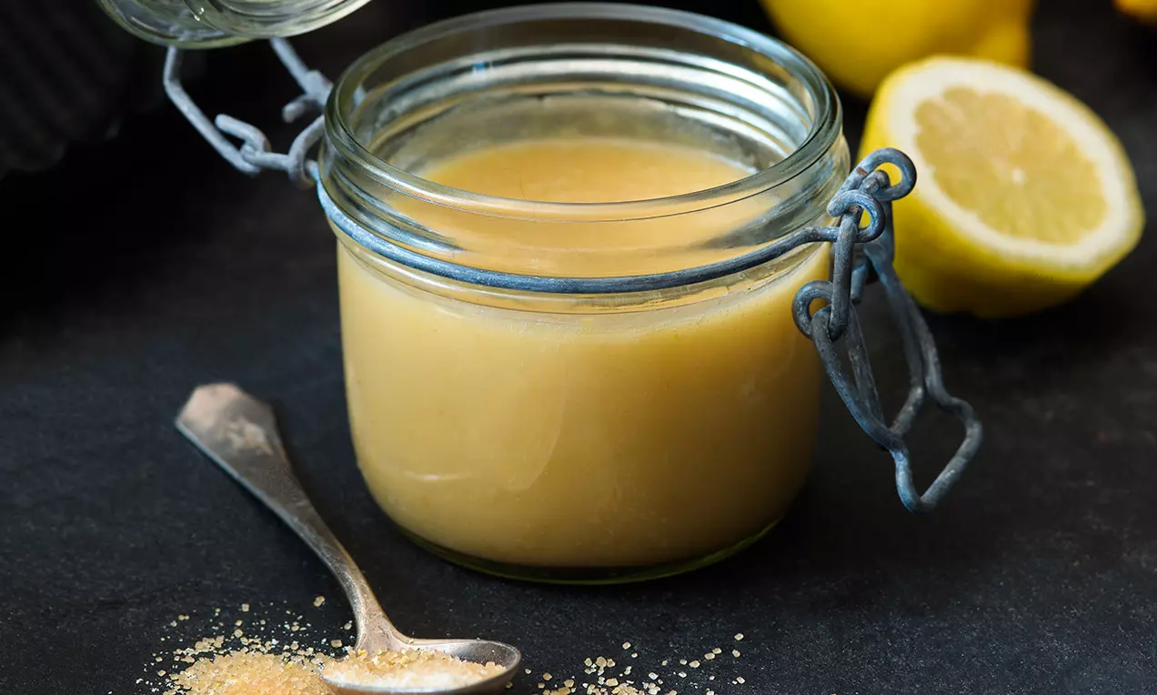 Světle žlutý krém hladké konzistence v průhledné sklenici s kovovým víkem, vedle kávová lžička a půlka citronu na tmavém povrchu