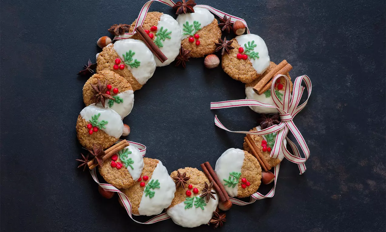 Kulaté perníkové cookies částečně polité bílou polevou, zdobené červenými bobulkami a zelenými lístky, uspořádané do věnce na tmavém pozadí