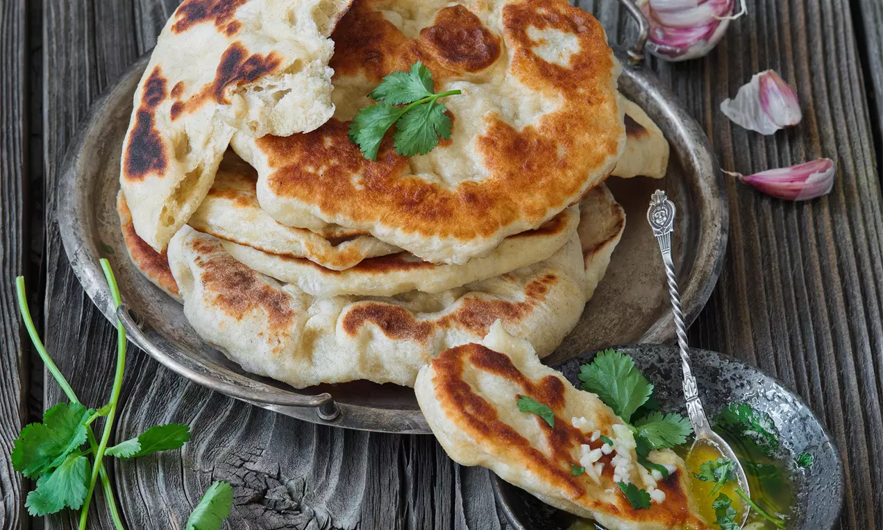 Zlatavé placky naan s lehkými hnědými opálenými skvrnami, měkká textura, vrstvené na kovovém talíři s čerstvým koriandrem a rozpuštěným máslem
