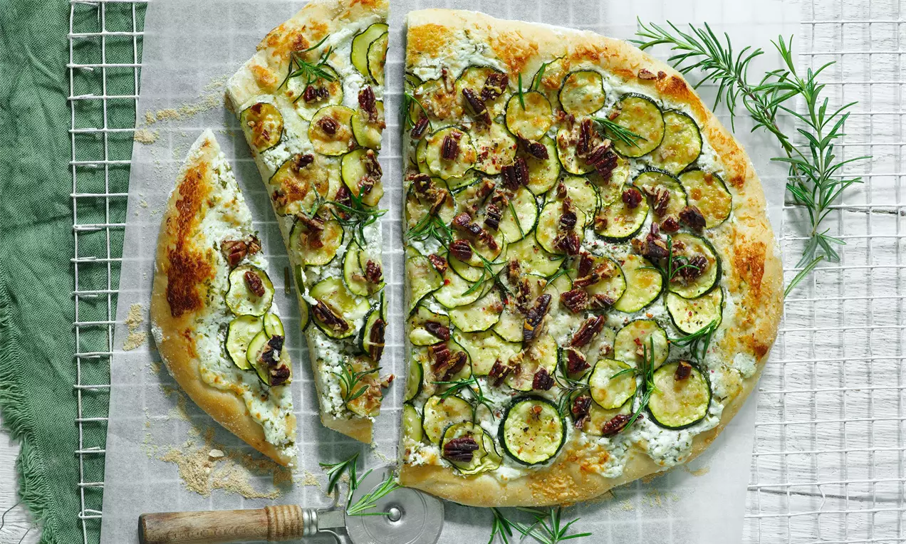 Cuketová pizza se zlatou křupavou kůrkou, zdobená tenkými kolečky cukety, sypaná bylinkami a ořechy na pečicím papíře s rozmarýnem