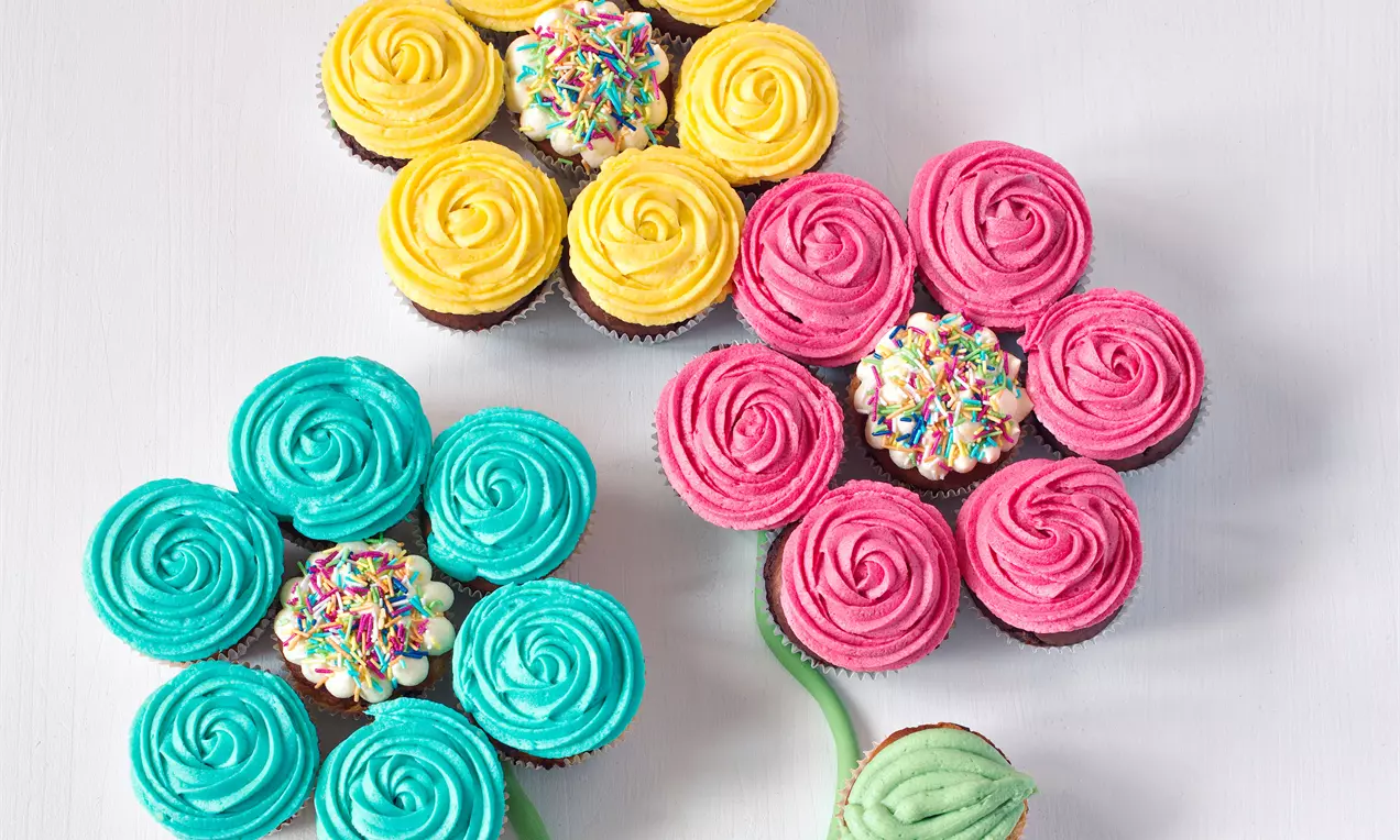 Muffiny zdobené barevným krémem do tvaru květů s růžovými, modrými a žlutými okvětními lístky a středem z posypky na bílém podkladu