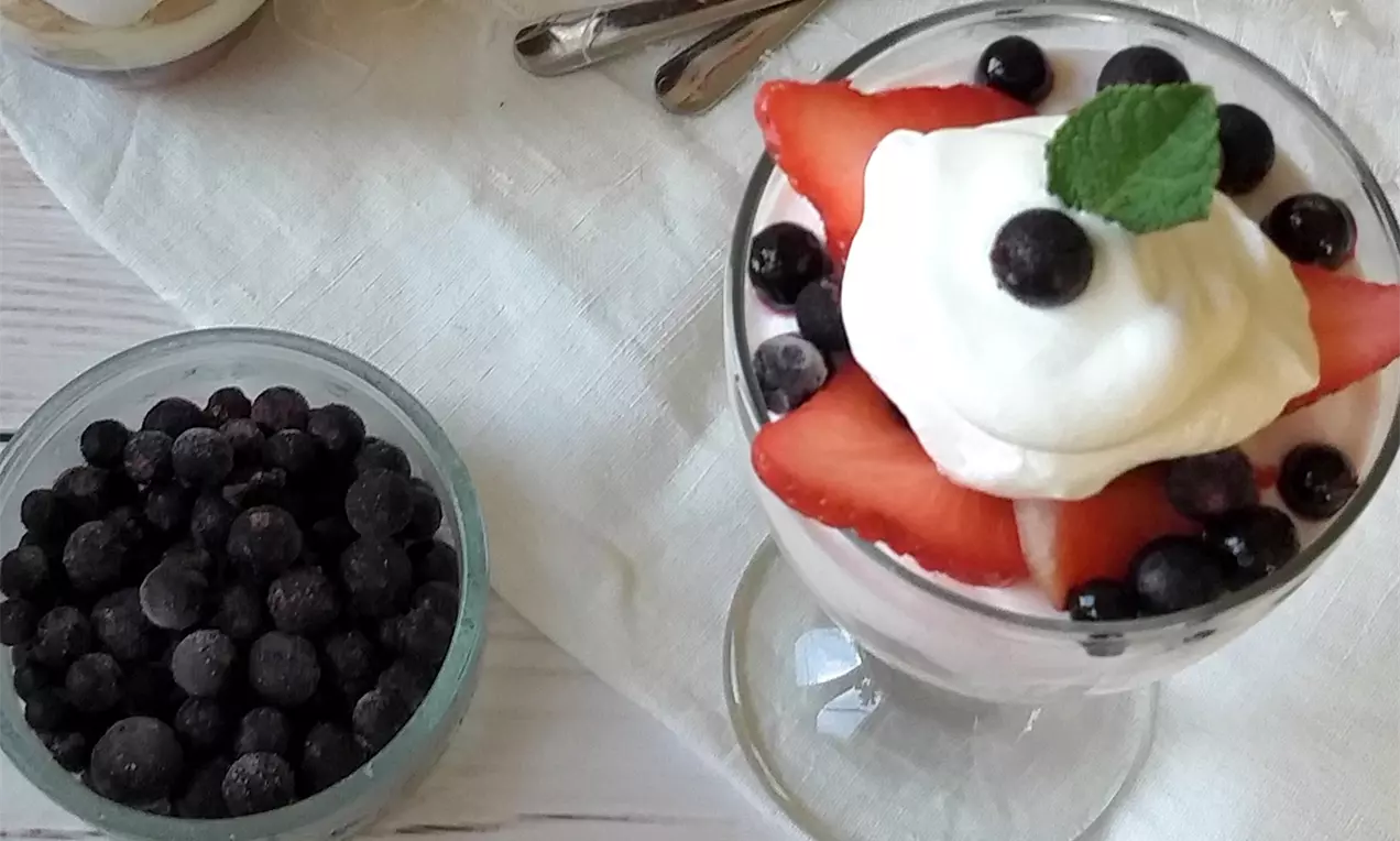 Sklenice s vrstveným trifle z jemného růžového krému, plátků jahod, borůvek a kopečku šlehačky, ozdobeného lístkem máty