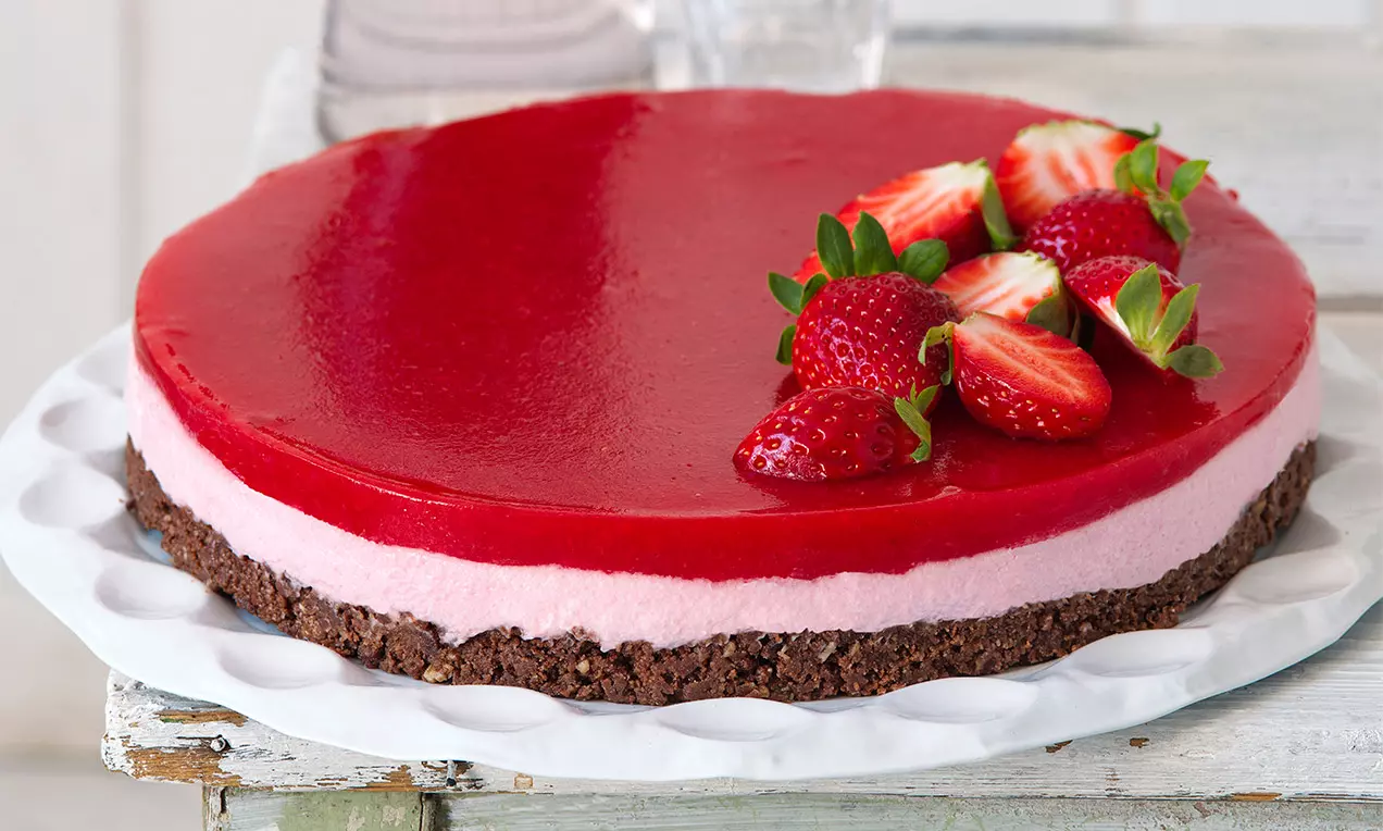 Kulatý jahodový cheesecake s červenou lesklou polevou, růžovým krémem a hnědým sušenkovým základem, ozdobený čerstvými jahodami na bílém talíři