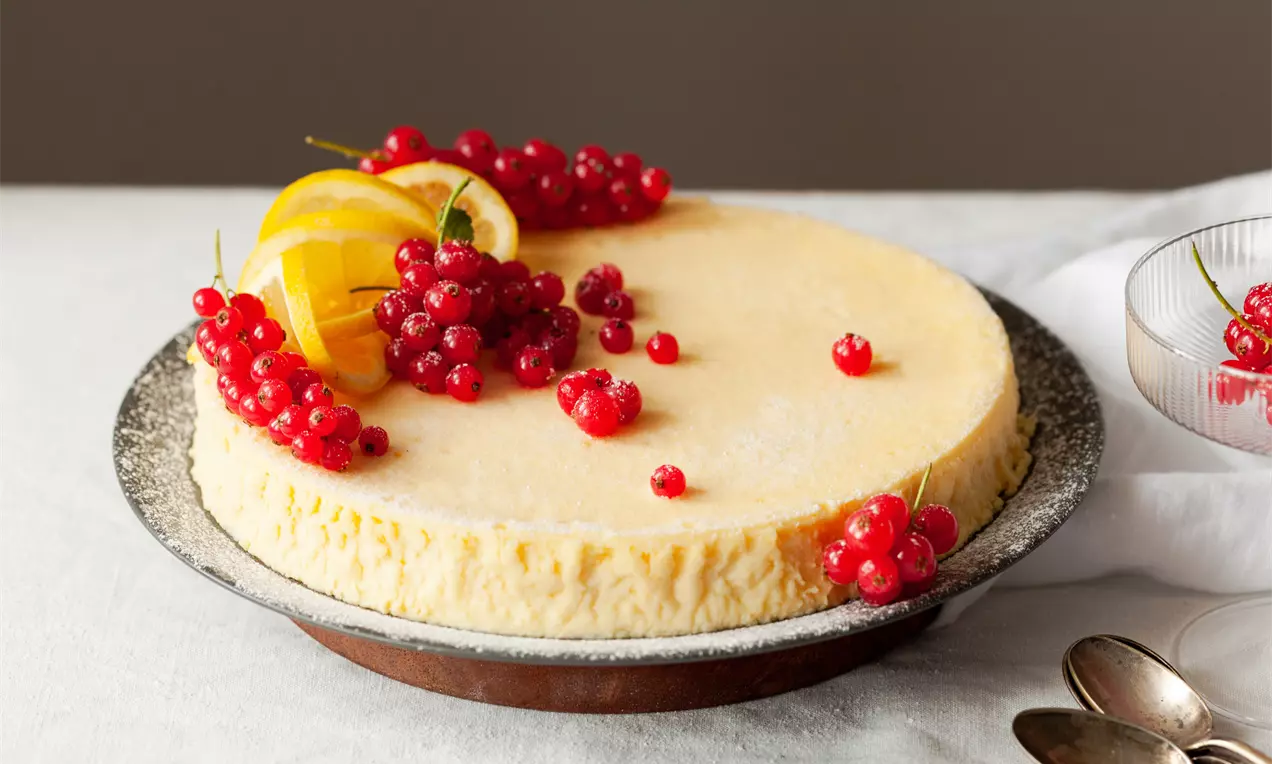 Světle žlutý ricottový koláč s hladkým povrchem, ozdobený červeným rybízem a plátky citronu na tmavém talíři s bílým ubrusem