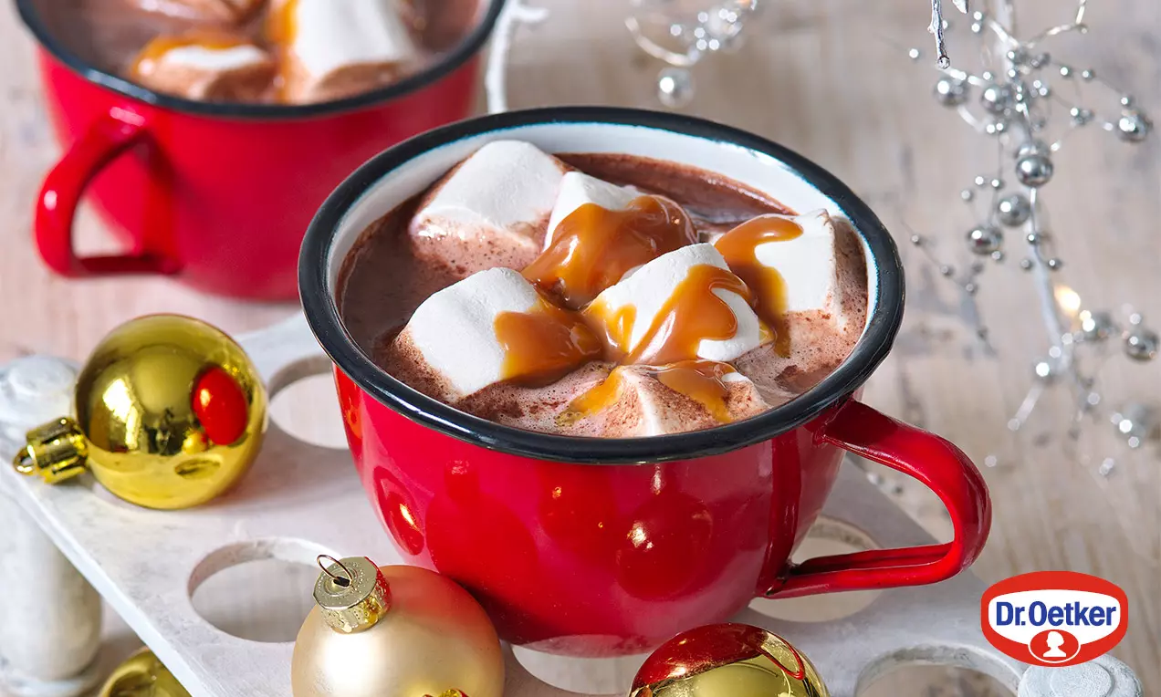 Horká čokoláda s pěnovou strukturou v červeném keramickém hrnku, zdobená bílými marshmallows přelitémi karamelovou polevou na dřevěném podnosu