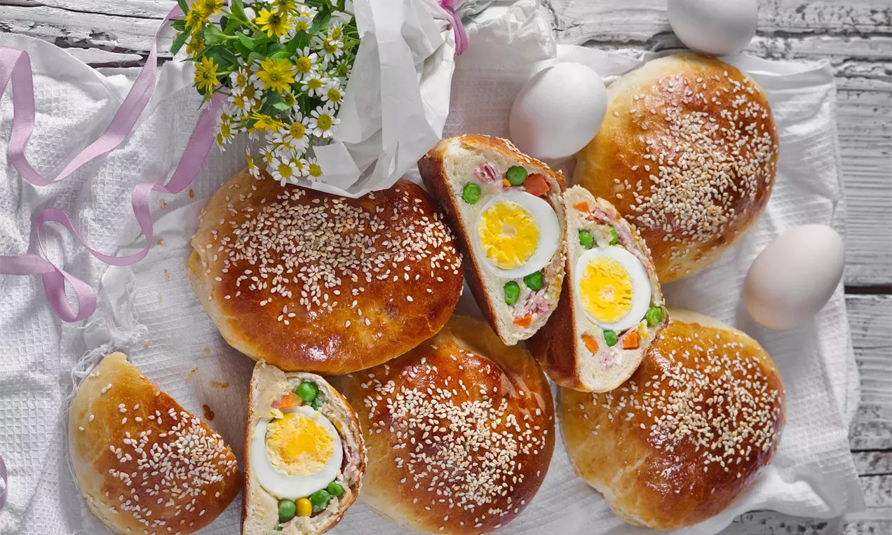 Zlatavé kulaté houstičky s lesklým povrchem posypaným sezamem, rozkrojené na půl s náplní z vajíčka, zeleniny a šunky na bílém ubrusu