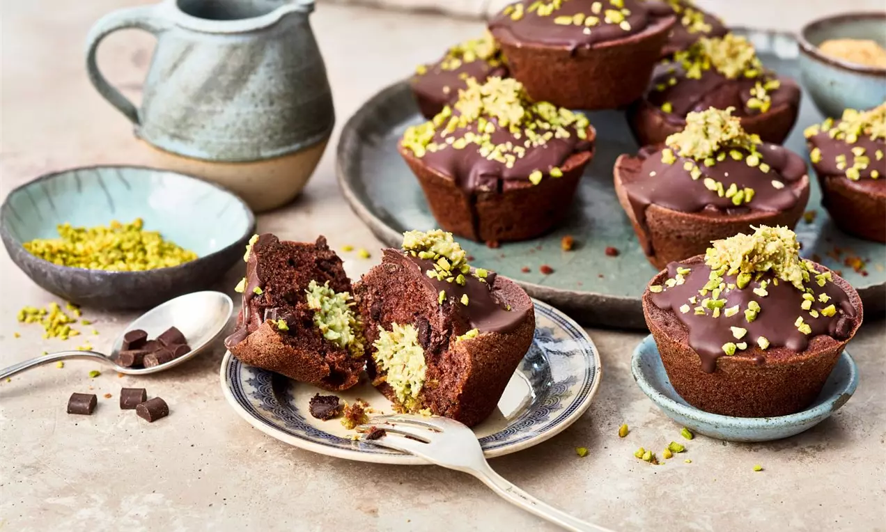 Čokoládové muffiny s lesklou polevou, posypané drcenými zelenými pistáciemi, uspořádané na keramickém talíři vedle nádobky s omáčkou