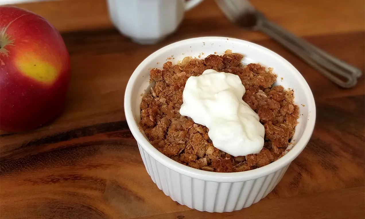 Jablečný crumble ve světlé keramické misce, zlatohnědá drobenka s hrubou strukturou, navrchu kopeček hladkého bílého krému na dřevěném stole