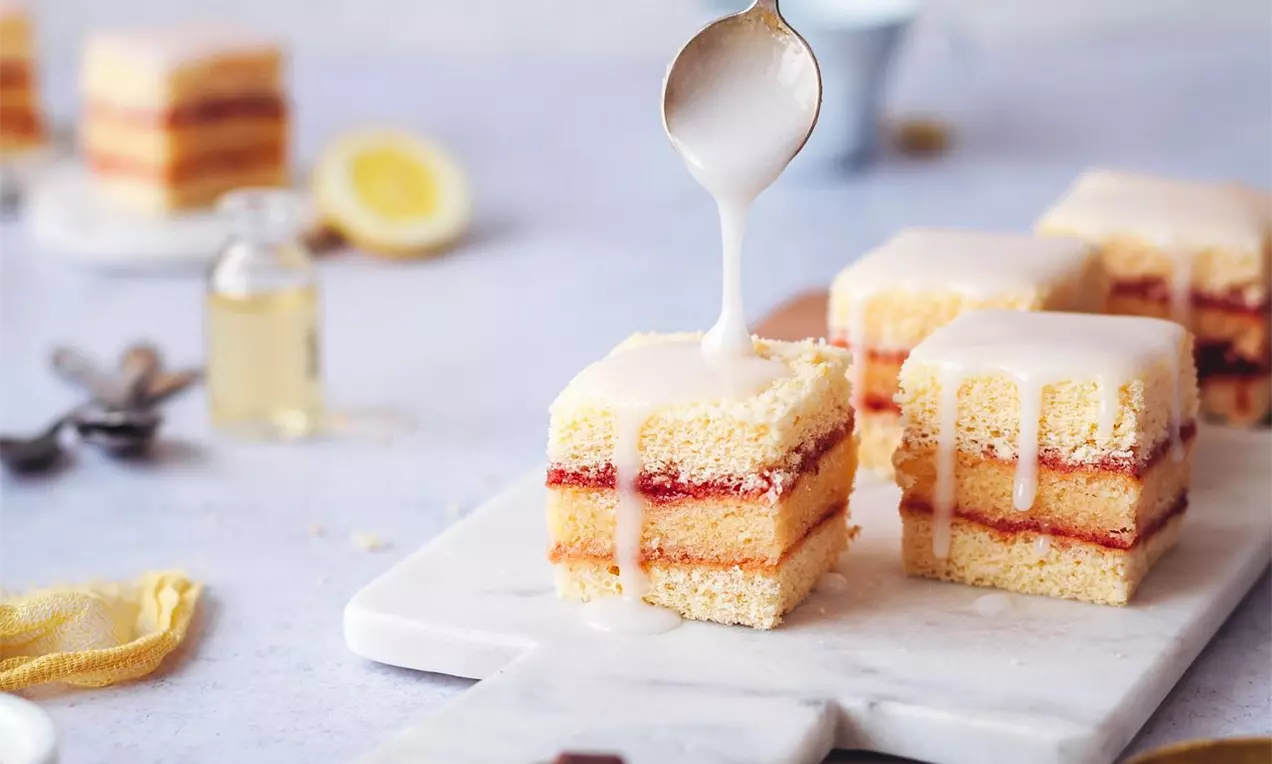 Čtvercové vrstvené řezy s jemným světlým piškotem, vrstvou červené marmelády a bílou citronovou polevou na mramorovém prkénku