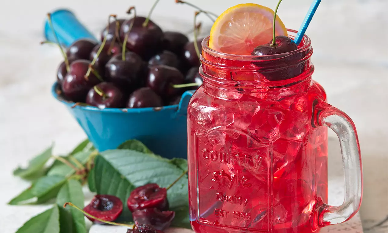Třešňově červený sirup s ledem v průsvitné sklenici, zdobený plátkem citronu, třešní a modrým brčkem vedle mísy čerstvých třešní