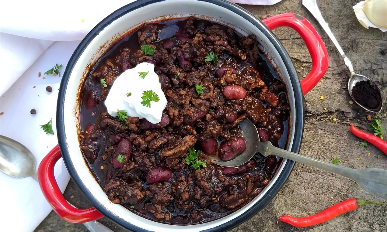 Tmavě hnědé chilli con carne s červenými fazolemi, zdobené kopečkem smetany a zelenou petrželkou v černo-bílé míse s červenými rukojeťmi