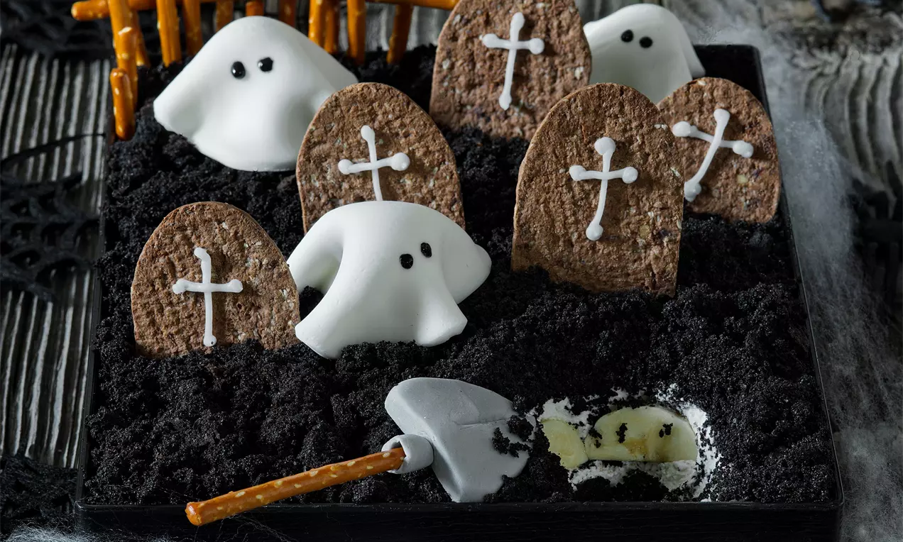 Halloweenský dort s černou drobenkovou vrstvou, čokoládovými náhrobky s bílými kříži, duchy z fondánu a lopatkou z sušenky