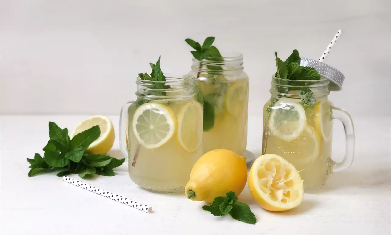 Světlá citronová limonáda v zavařovacích sklenicích, plátky citronu, čerstvá máta, dekorace citronů a lístků na světlém podkladu