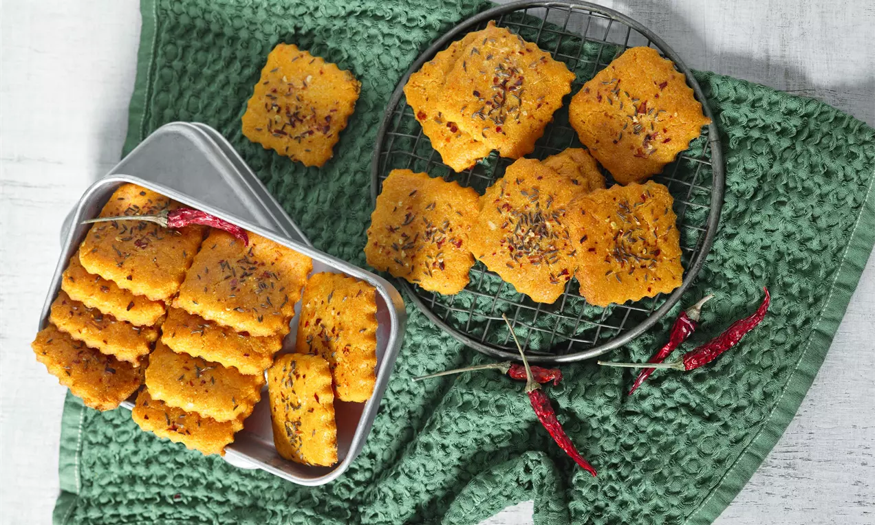 Křupavé čtvercové chlebíčky zlatavé barvy posypané drobnými chilli vločkami, uspořádané na mřížce a v plechové krabičce na zeleném látkovém podkladu