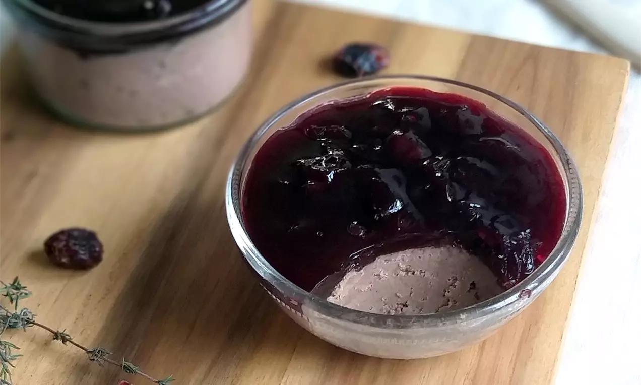 Krémová světle hnědá krůtí paštika ve skleněné misce, pokrytá tmavě červenými brusinkami v lesklém vinném želé, na dřevěném prkénku