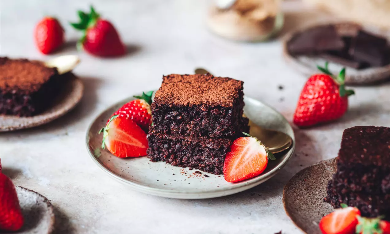 Dva silné čtverce tmavého brownies s jemnou drobivou strukturou na světlém talíři, doplněné čerstvými červenými jahodami