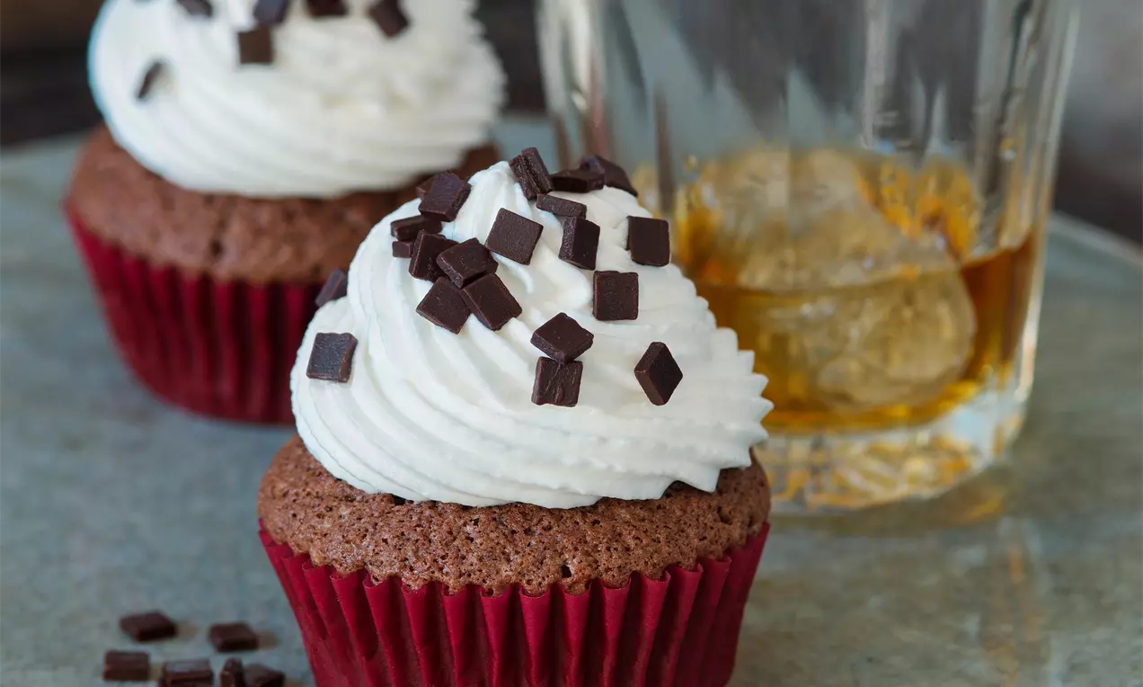 Čokoládové muffiny v červených papírových košíčcích s bílou šlehačkou, posypané čokoládovými kostičkami a sklenicí whisky v pozadí