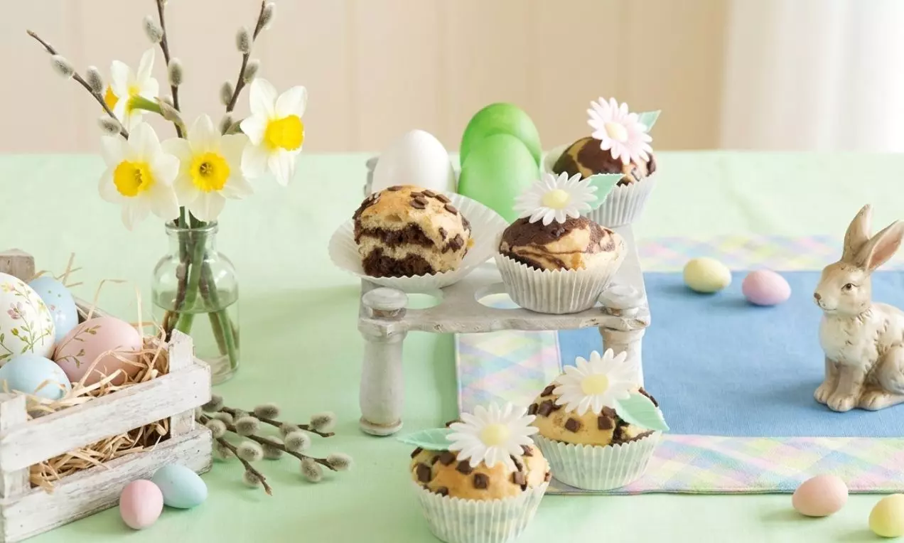 Dekorativní muffiny mramorovaného vzhledu v papírových košíčcích, zdobené bílými květy se zelenými lístky na pastelově laděném stole, AI-labeled image