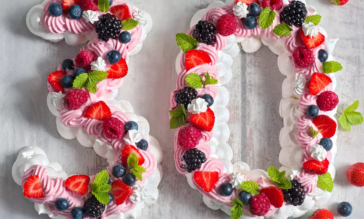 Bílá pavlova ve tvaru číslice 30 zdobená růžovým krémem, čerstvými malinami, jahodami, borůvkami, ostružinami, lístky máty a květy