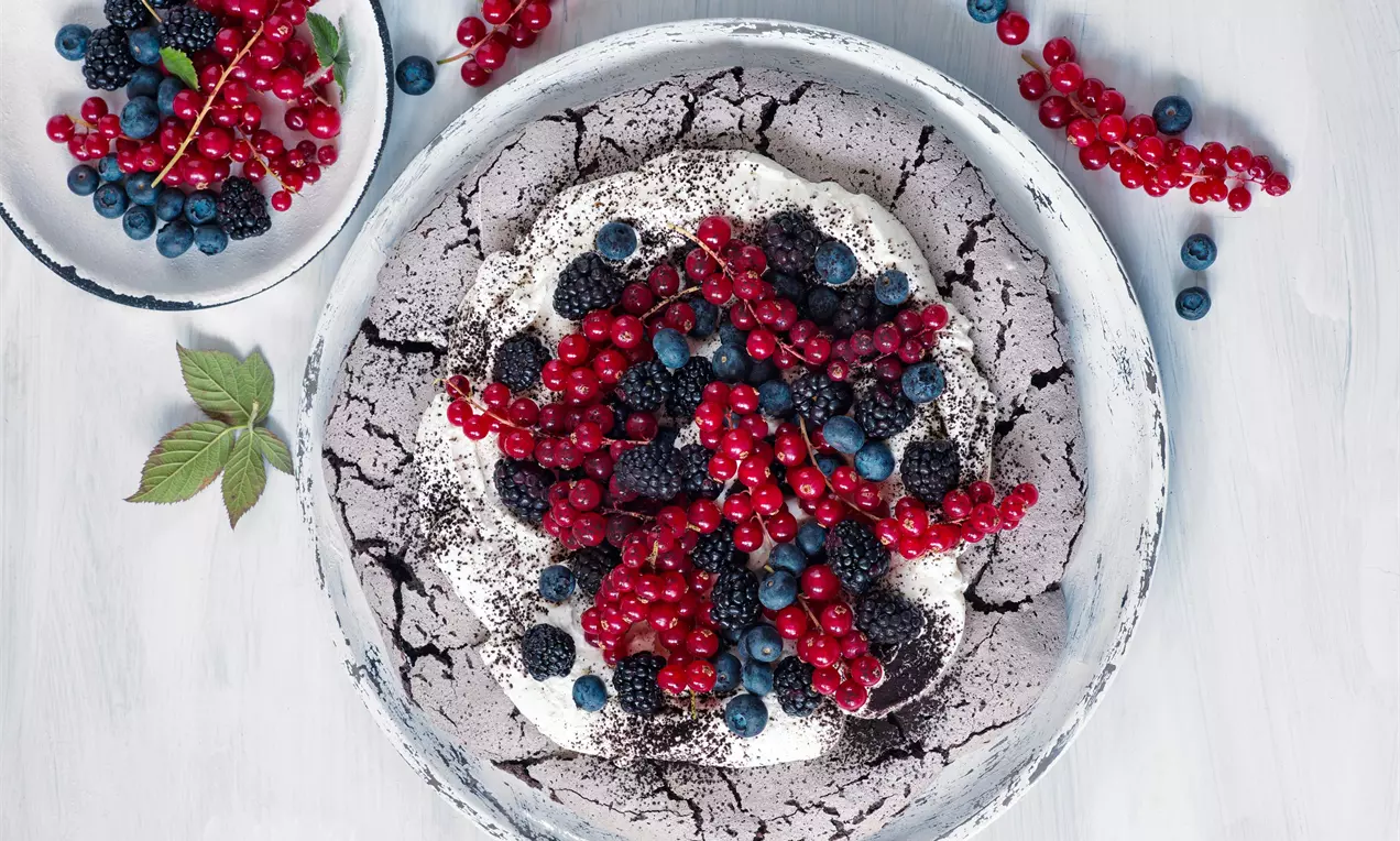 Pavlova s popraskaným černým kakaovým základem, nadýchanou bílou pěnou zdobenou lesklými červenými ríbezlemi, ostružinami a borůvkami