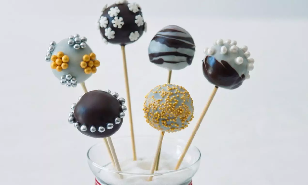 Čokoládové a polevou potažené kulaté cake pops na dřevěných tyčinkách, zdobené perličkami, vločkami a zlatými i stříbrnými akcenty ve sklenici