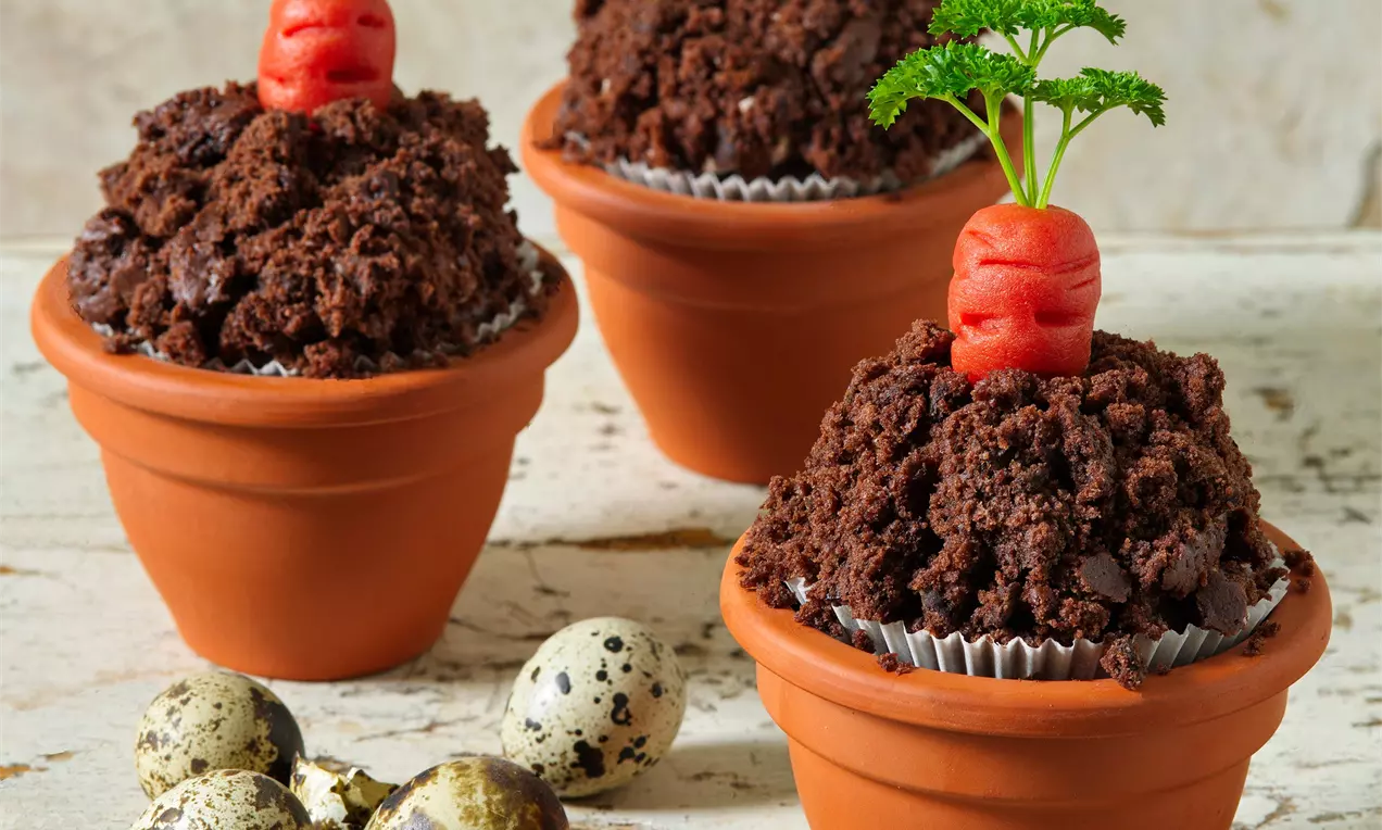 Čokoládové muffiny s drobivou texturou ozdobené červenou marcipánovou mrkvičkou s zeleným lístkovím, podávané v malých terakotových květináčích