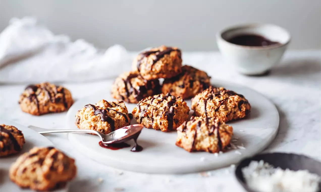 Zlatavé ovesné cookies nepravidelného tvaru na mramorovém podnosu, s tmavou čokoládovou polevou, lžičkou a miskou čokoládové omáčky