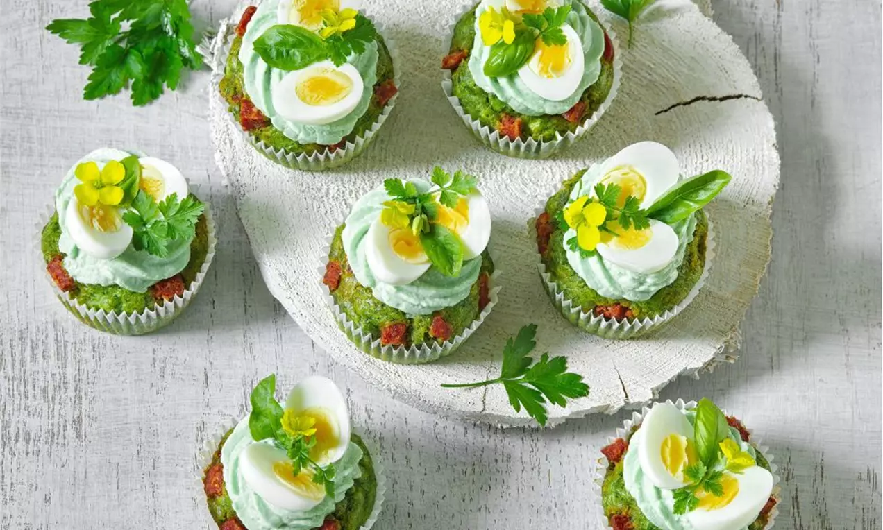 Špenátové cupcakes s jasně zeleným těstem, čepicí z bílé šlehané polevy, zdobené polovinou vajíčka, bylinkami a kvítky na světlém talíři